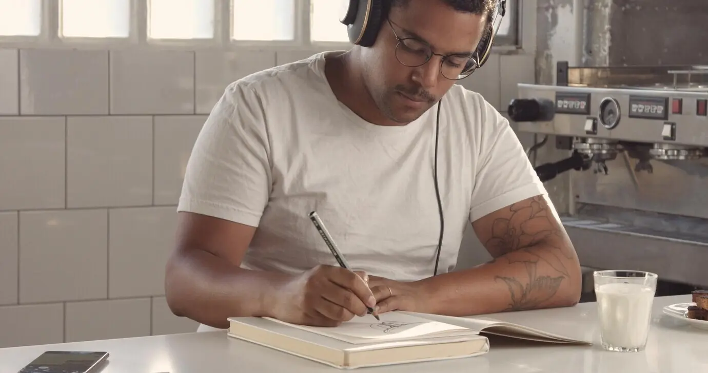 Ein Mann gemischter ethnischer Herkunft zeichnet in einem sonnigen Loft und hört über Ohrhörer Musik; Bilder mit Sonnenreflexen.